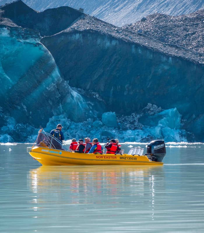 Yellow boat carrying tourists with life jackets near blue glacier ice on calm water.