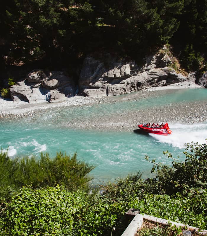Red jet boat speeding through a clear turquoise river surrounded by trees and rocks.