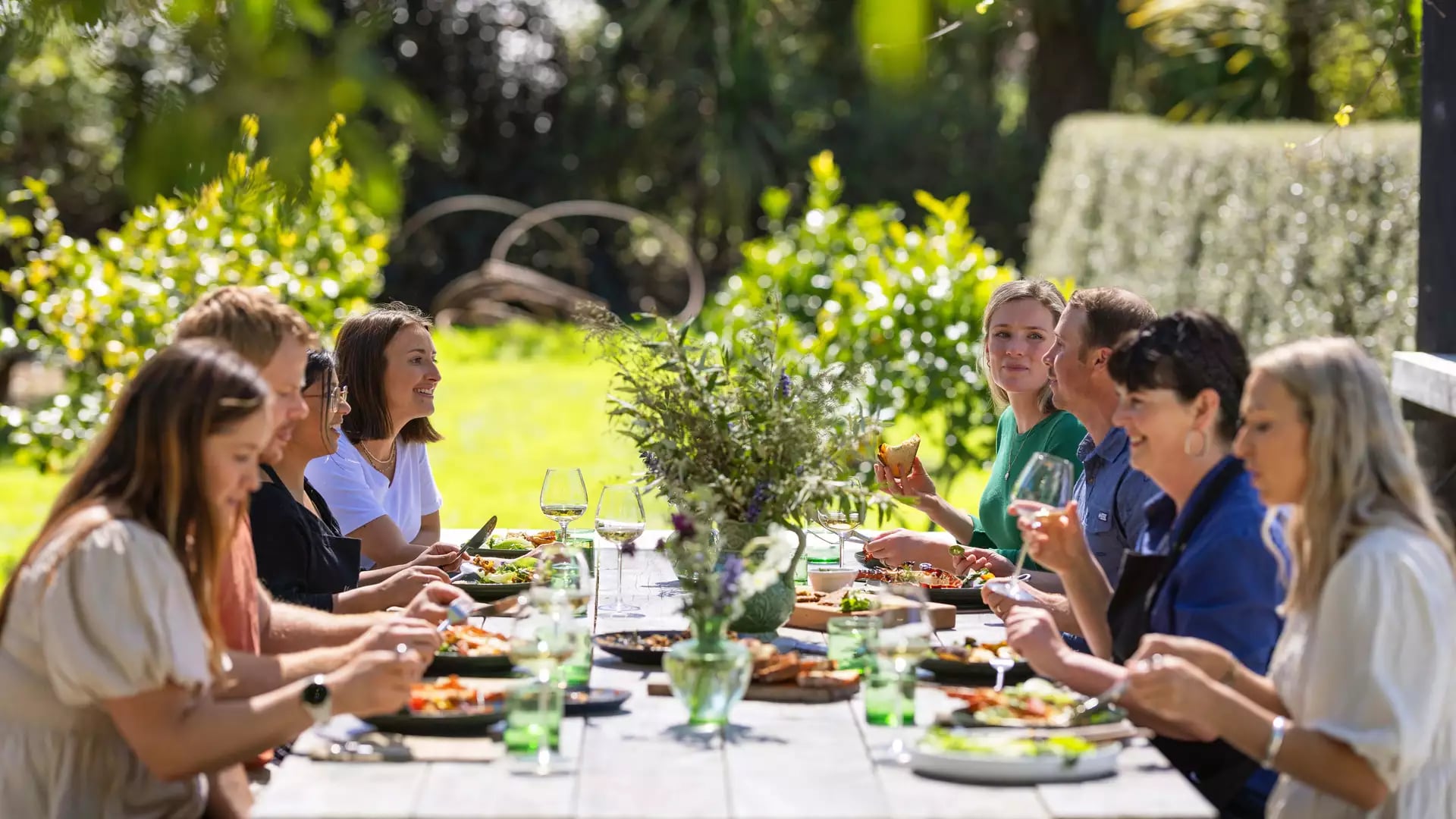 Group of people eating lunch outdoors at a long table with plants and glasses in daylight.