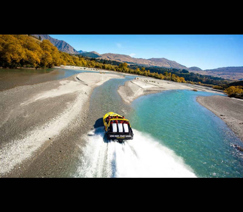 An aerial view of a yellow and black jet boat speeding through a shallow, turquoise braided river, leaving a white wake, surrounded by golden autumn trees and rugged mountains under a clear blue sky. Credit: KJet