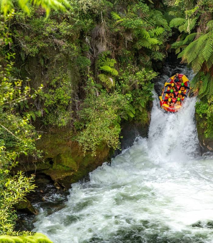 A red white-water raft filled with people in yellow helmets plunges down a steep 7-metre waterfall surrounded by lush green native ferns and forest.