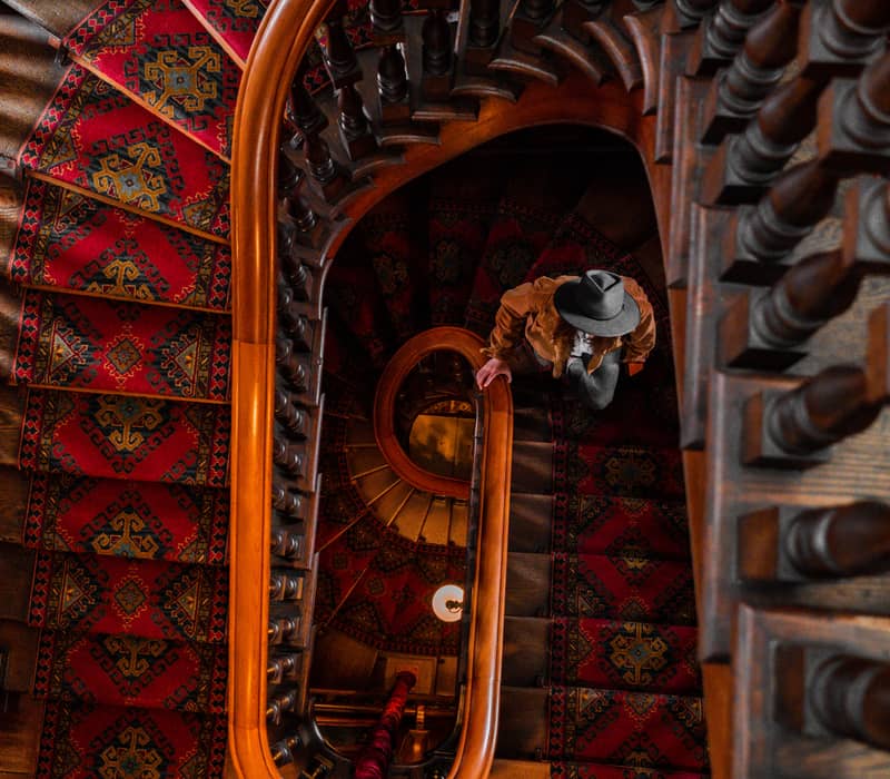 Top view of a spiral staircase with red carpet and wooden railing, a person wearing a hat is descending.