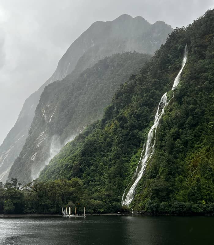 Waterfall pouring down a green mountain into a lake with mist and a wooden dock.