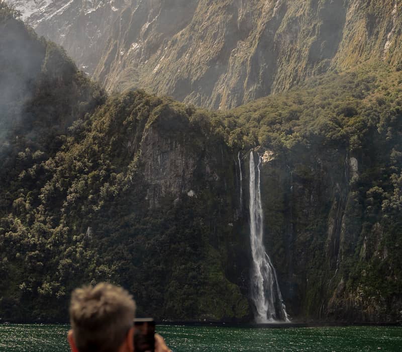 Person taking photo of waterfall cascading down mountains next to a green lake