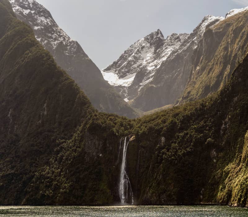 Tall waterfall flowing into lake with steep forest-covered cliffs and snow-capped mountains behind.