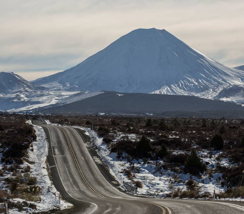A massive, snow-covered conical volcano dominates the horizon at the end of a long, winding road through a wintry volcanic landscape under a pale sky.