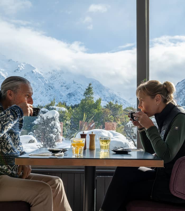Couple drinking coffee inside a cafe with snow-covered mountains seen through the window.