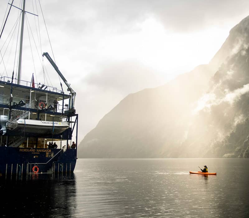 Ship Fiordland Navigator anchored in foggy Doubtful Sound with kayaker on calm water near mountains.