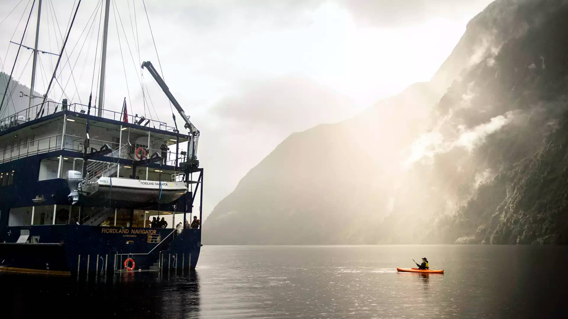 Ship Fiordland Navigator anchored in foggy Doubtful Sound with kayaker on calm water near mountains.