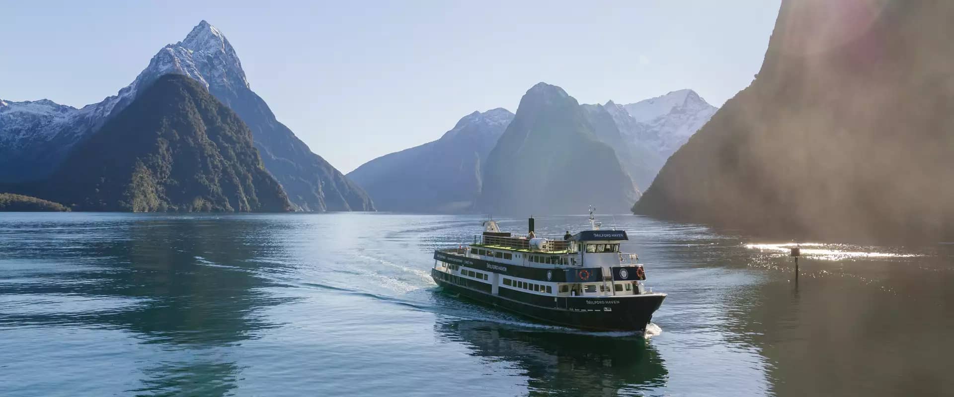 Milford Mariner cruise ship sails in misty fjord surrounded by tall mountains and cloudy skies.