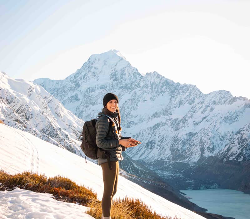 Woman standing on a snowy slope with backpack overlooking a lake and snow-covered mountains.