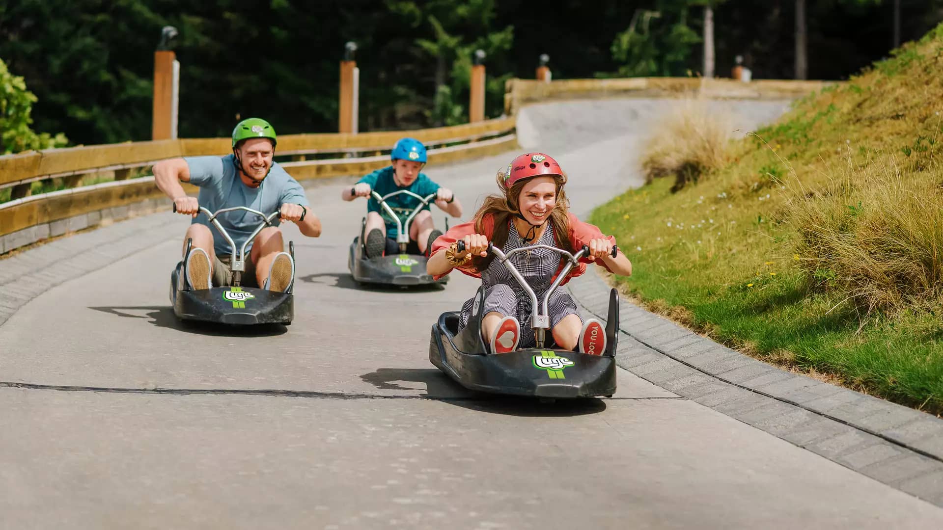 Three people race down the Skyline Queenstown Luge track in colorful carts, with Lake Wakatipu and the snow-capped Remarkables in the background.