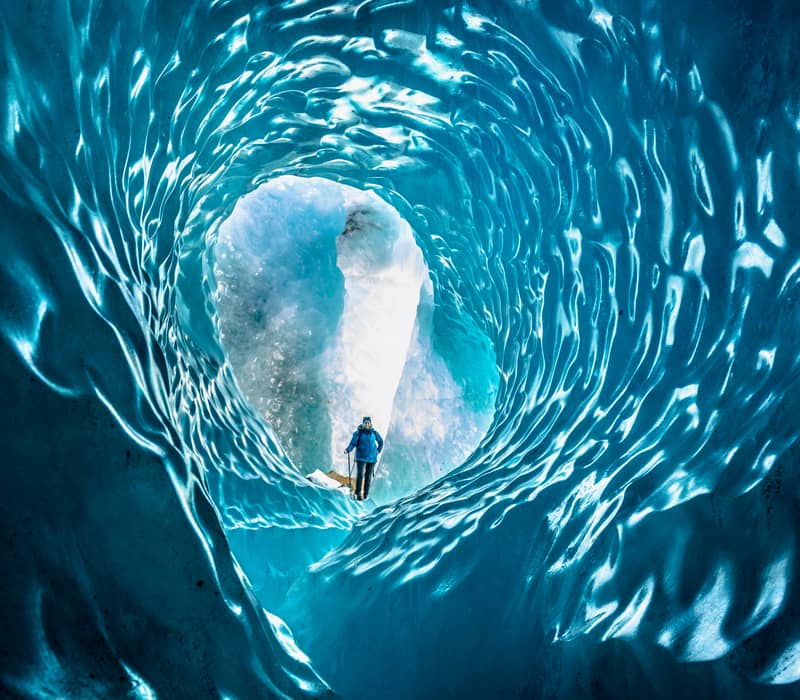 Person standing inside a blue ice cave with textured glacier walls.