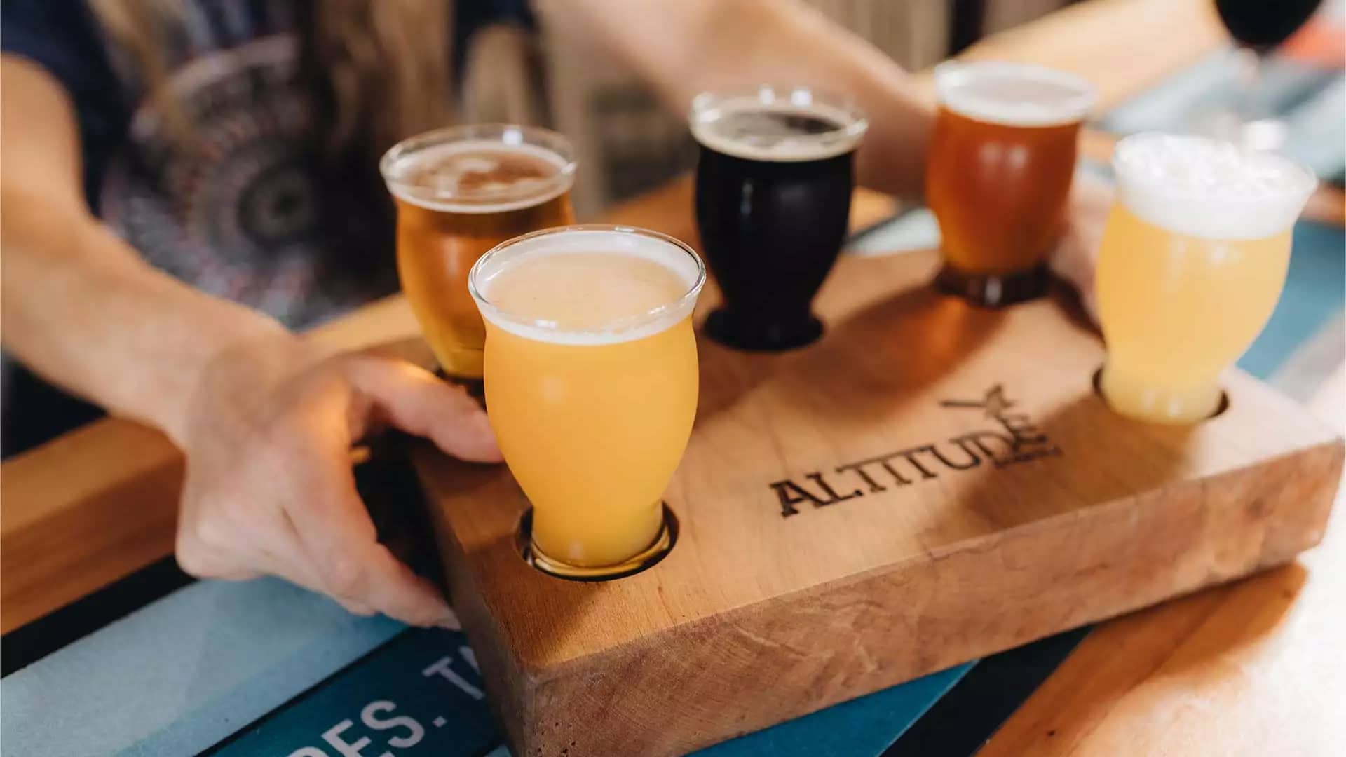 Hand holding wooden tray with six glasses of craft beer in diverse colors for tasting.