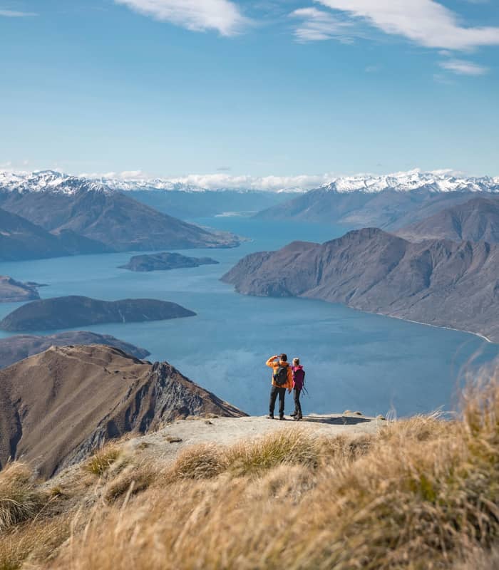 Two hikers on Roy's Peak with a wide view of Lake Wanaka and snowy mountains in the background.