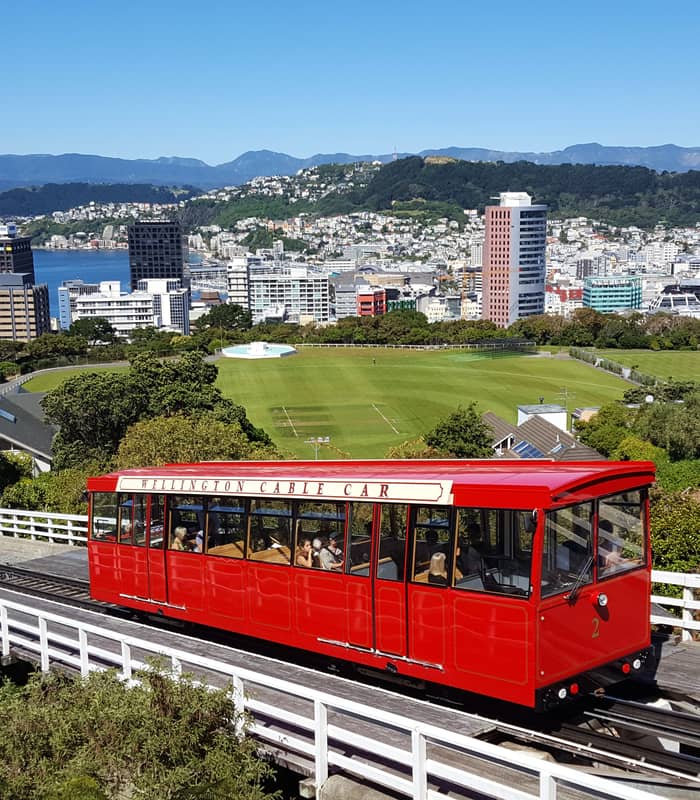 The bright red Wellington Cable Car travels down its tracks overlooking Wellington city, the harbor, and the surrounding hills under a clear blue sky.