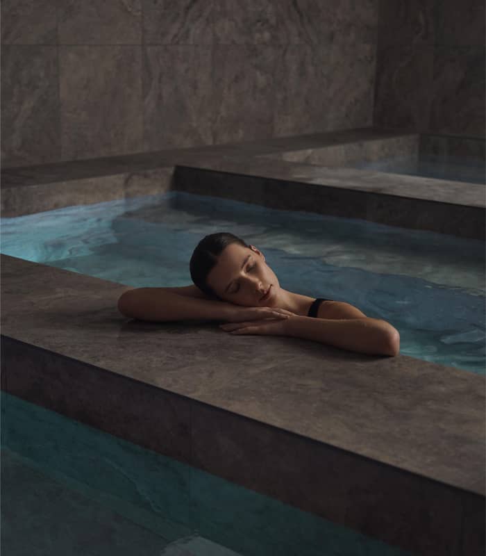 Woman resting her head on her arms at the edge of an indoor marble bathhouse pool.