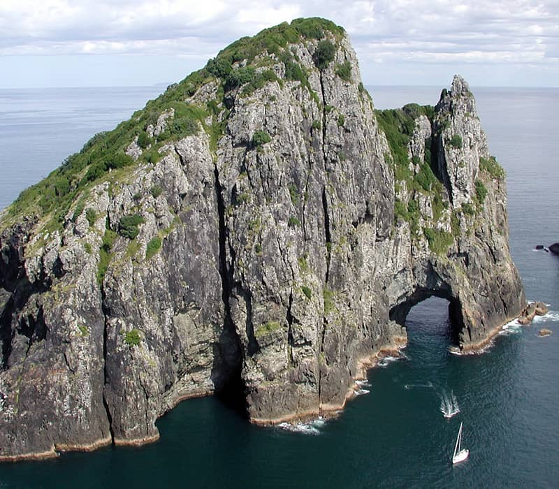 Large rocky island with natural arch in the ocean and small sailing boats nearby.