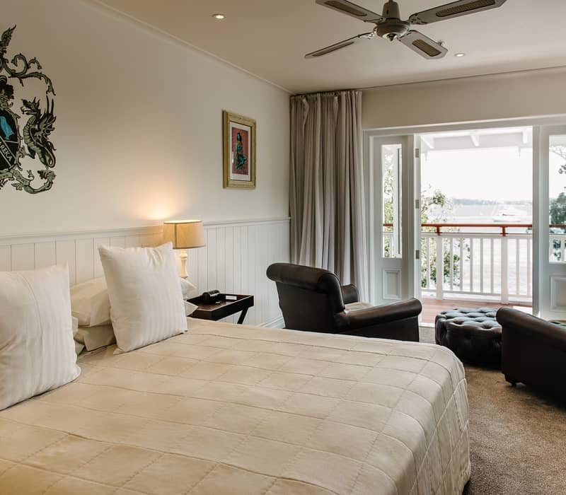 Hotel room with bed, armchairs, and balcony overlooking waterfront with boat.