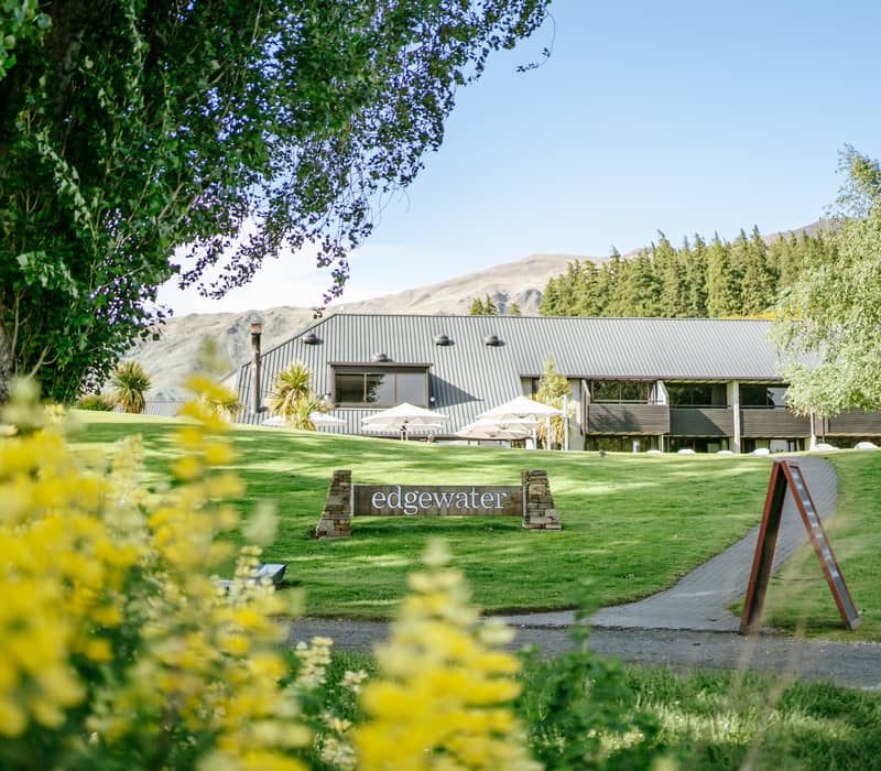 Edgewater hotel building with green lawn, yellow flowers, trees, and mountains in the background.