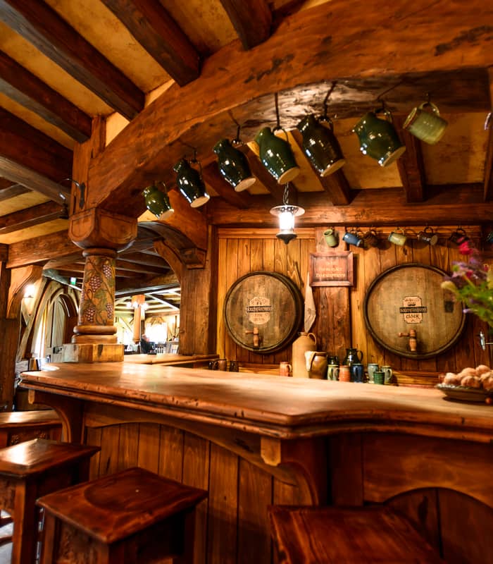 Rustic wooden pub bar with stools, hanging mugs, barrels, and flower arrangement.