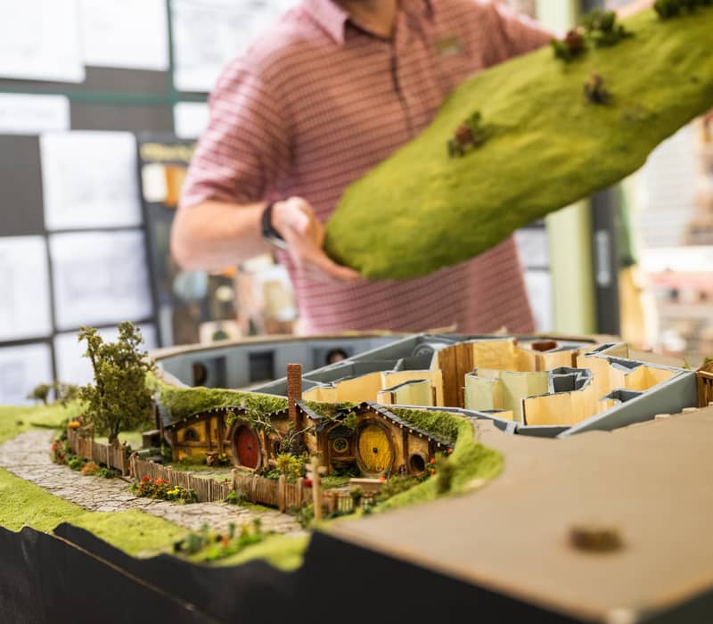 Person holding a section of a detailed Hobbiton scale model with miniature houses and greenery.