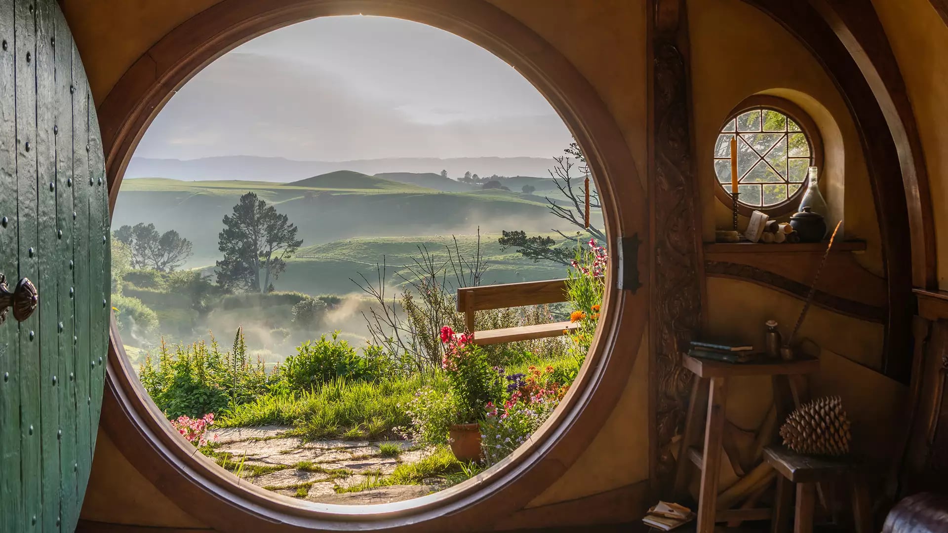 Inside view of a hobbit hole with a round door open to a green garden and rolling hills beyond.