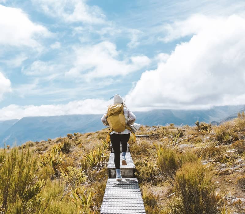 Person hiking on a boardwalk trail in the mountains with a yellow backpack and cloudy sky.
