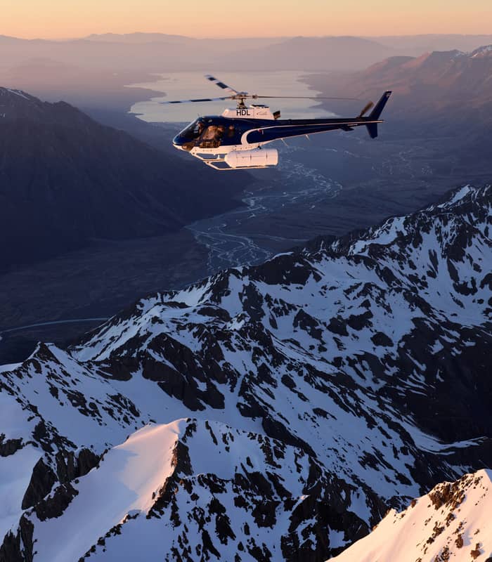 Helicopter flying over snowy mountains and glaciers at sunset with river and lake below.