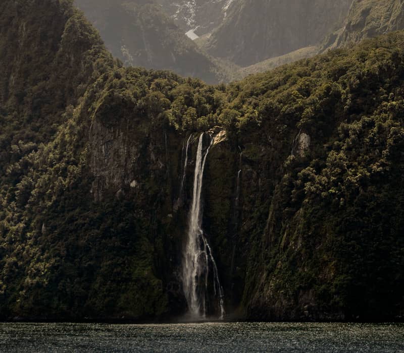Waterfall flows down forested cliff with snowy mountain peaks behind and water in the foreground.