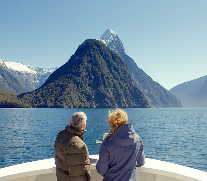 Two people on a boat looking at a snow-capped mountain over a blue lake on a clear day.