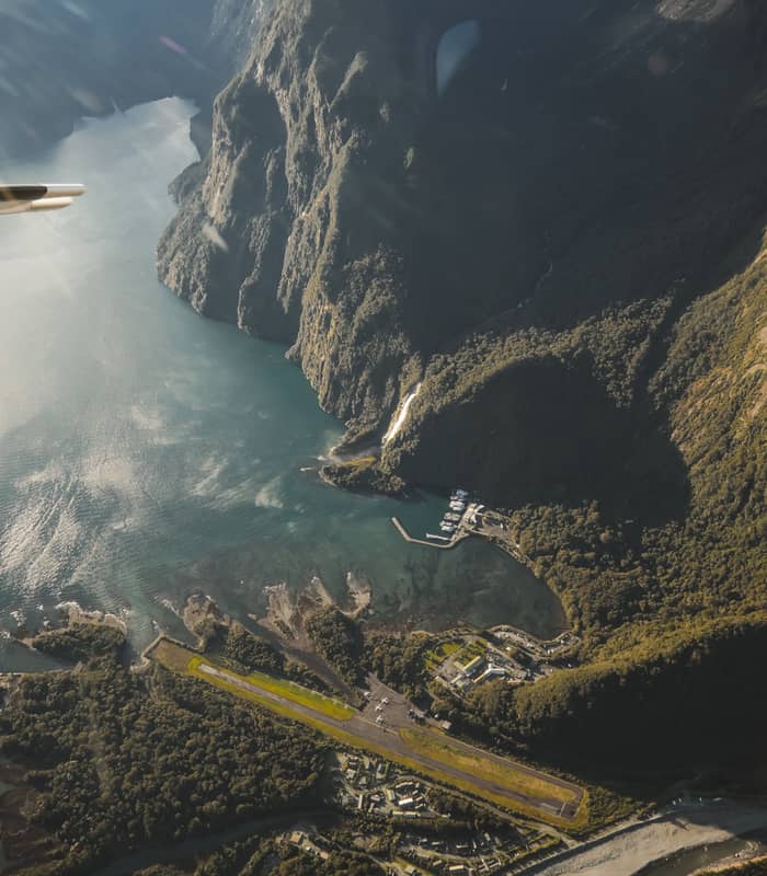 Aerial shot of Milford Sound showing mountains, runway, waterfalls, harbor, and river.