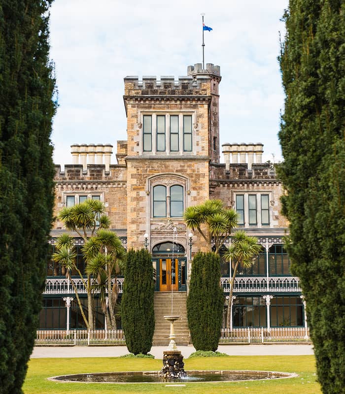 Larnach Castle viewed through tall hedges with a fountain and garden in front.