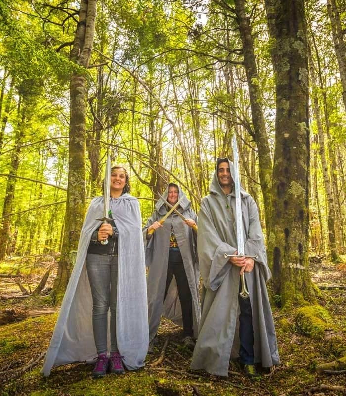 Three people wearing capes and holding swords pose in a bright green forest.