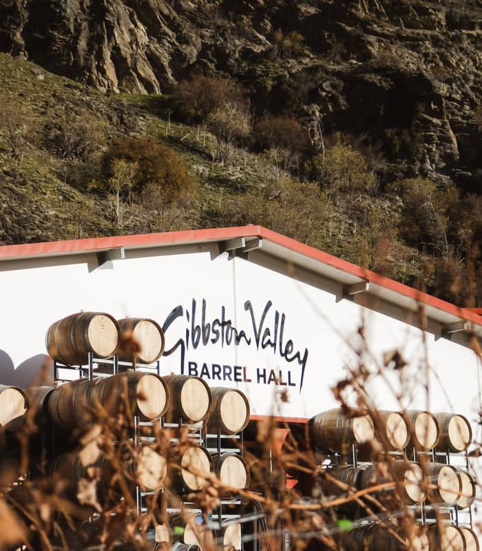 Wooden wine barrels stacked outdoors at Gibbston Valley Barrel Hall with hillside in background.