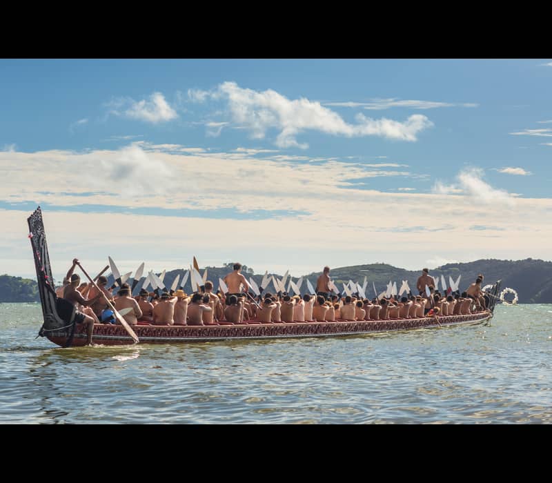 People paddling a traditional waka canoe on water with hills and blue sky in background.