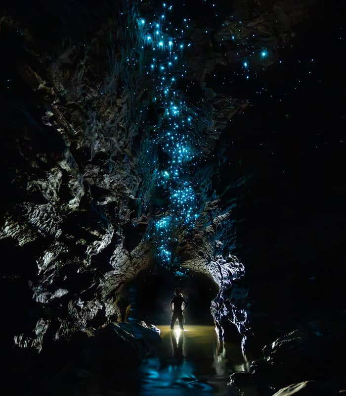 Silhouette of person standing inside a dark glowworm cave with blue glowing lights on the ceiling and reflections in the water.